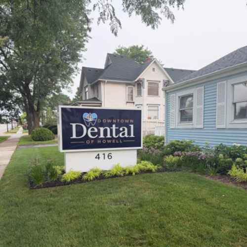 Dental office sign for Downtown Dental of Howell, number 416, on a landscaped lawn with a blue house nearby.